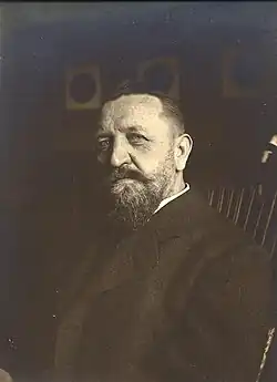 A dark-bearded man in a dark suit sitting in a chair