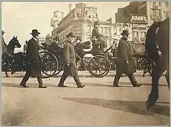 Several men walk down the road. Roosevelt sits in a horse-drawn carriage. A large crowd watches in the distance.