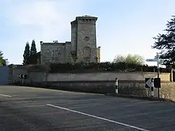 Stone building with tower to right hand side. In front is a wall separating the building from the road.