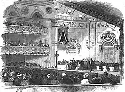 The interior of an elegant concert hall, an elegant audience, and on the stage, many men participating in the ceremony.
