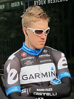 A man in his late twenties wearing a black and white cycling jersey with blue trim, and sun shades.