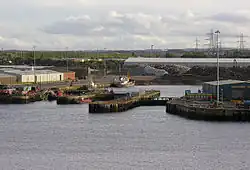 A view of a small dock as seen from a departing ferry. Industrial buildings surround, and a small boat is docked in the corner.