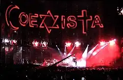 A view of the stage being filmed, with "COEXISTA" spelled out on the video screen, using an Islamic crescent as a "C", a Star of David as an "X", and a Christian cross at a "T". A camera in the middle shows the concert being filmed.