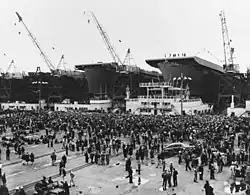 Black and white photograph of numerous people standing on a concrete surface, in front of three ships under construction with cranes on top of them