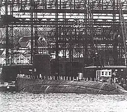 An incomplete submarine lays a rest; tall steel gantries and a dock dominate the background.