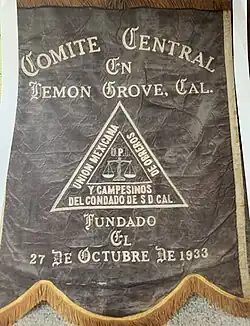 A banner type flag with black background and gold writing with a triangular logo in the center stating the name of the Union, Union Mexicana De Obreros Y Campensinos Del Condado De S.D.Cal and a justice scale in the center. States founded on the 27th of October 1933.