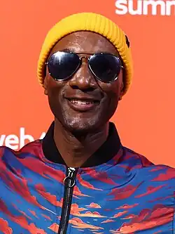 close-up of Aloe Blacc standing in front of a bright orange background, wearing a patterned jacket, yellow beanie, and dark aviator sunglasses, grinning at camera