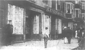 A black and white photograph of a storefront, with boarded windows, and some individuals outside on the sidewalk