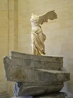 A marble sculpture of the Winged Victory of Samothrace, a headless and armless figure with large wings, standing on a stone base resembling the prow of a ship. The statue's flowing drapery and dynamic pose suggest movement, as though caught in the wind. It is displayed indoors against a beige stone wall.