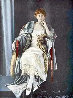Colour photo of middle-aged white woman, expensively dressed, with dark reddish hair, seated with head resting on one hand, looking at camera