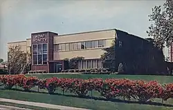 An exterior view of a two-story modernist structure with landscaping out front. The building is a buff color with an offset maroon-clad box. On top of the box are large silver letters on two lines, "W D A F" and "W D A F - T V".