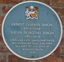A circular plaque coloured blue mounted on a brick wall, containing the coat of arms of Manchester City Council at the top, the white text "Ernest Darwin Simon (1879–1960)" and "Shena Dorothy Simon (1883–1972)" in the centre, followed by "Lord and Lady Simon purchased Wythenshawe Hall and Park as a gift to the City of Manchester in 1926"