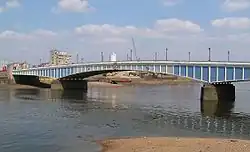 A long low bridge across a wide river. The bridge is painted in varying shades of blue, which render it hard to see against the water and sky.
