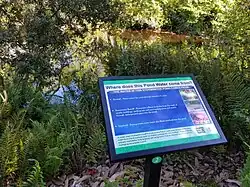 A sign about water in front of a pond with plenty of surrounding plants and foliage.