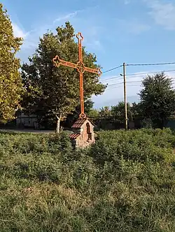 A large, orange-painted metal Orthodox cross mounted on a small stone and brick base. It stands in a field of overgrown green bushes, with trees in the background.