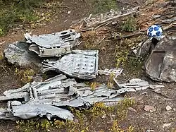 A yellow jersey with “12” and “SRS” hangs from a tree trunk at a memorial site. A white cross is planted nearby, and fallen logs and scattered vegetation cover the sloped, forested area. A piece of dark clothing hangs over one of the logs in the background.