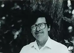A man with dark hair, large square glasses, and a white collared shirt is shown with a small smile