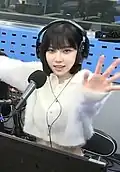Woman with a short black bob hairstyle wearing headphones and speaking into a microphone in a radio studio. She is dressed in a white, fuzzy, cropped cardigan and is smiling brightly while extending her arms toward the camera.