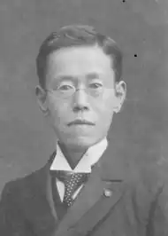 Black-and-white photograph of a young Japanese man with round glasses, wearing a morning suit.