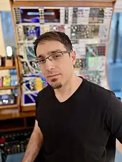 Andrew Aversa wearing a black v-neck t-shirt, standing in front of a modular synthesizer in his home studio, looking directly at camera with slight squint, head tilted to the right