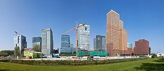 Photo of nine building towers in varied colors and a construction site with in the center a yellow tower crane, in the background on the left is a second tower crane